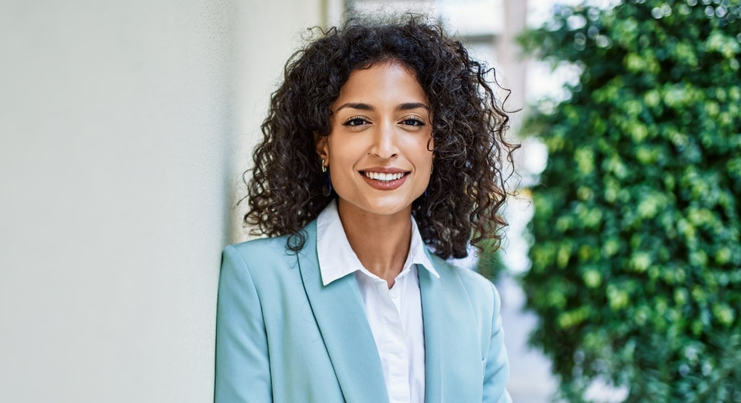 A woman in a light blue blazer smiles while standing outside near a wall, with greenery in the background.