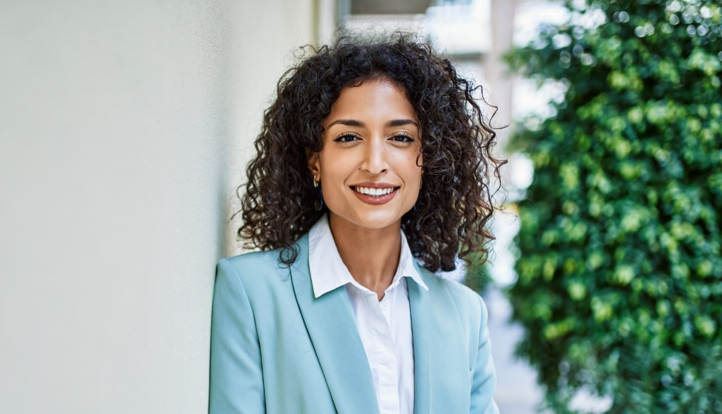 A woman in a light blue blazer smiles while standing outside near a wall, with greenery in the background.